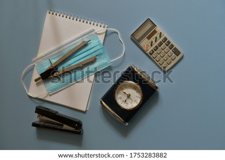Back to school concept with gray divider, blue medical mask (protective mask), white notebook, dark alarm clock, black stapler and light gray calculator on a blue background