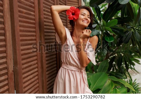 Excited Asian model enjoying tropical vacations.  Wearing pink dress. Hibiscus flower in hairs.