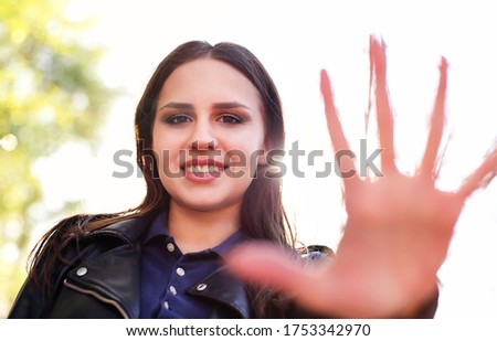 Content young female in leather jacket showing palm at camera and smiling against bright sunlight in park