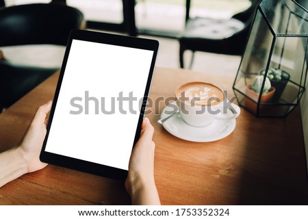 A woman hands are holding a mockup of black tablet with blank white screen   in the cafe with a white cup of latte coffee. sitting on the wood table, clipping path included Royalty-Free Stock Photo #1753352324