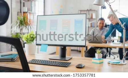Desktop Computer Standing on the Desk in the Modern Creative Office. Screen Showing Smartphone Application UI / UX.  In the Background Bright Studio with Team of Young Professionals Work on Computers.