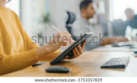 Anonymous Young Woman Sitting at Her Desk She's Drawing, Writing and Using Pen with Digital Tablet Computer. Focus on Hands with Pen. Bright Office where Diverse Team of Young Professionals Work.