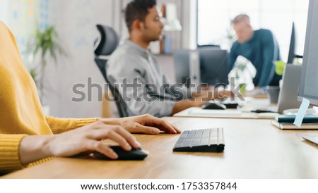 Portrait of Anonymous Creative Young Woman Sitting at Her Desk Using Computer Mouse and Keyboard. In the Background Bright Office where Diverse Team of Young Professionals Work.