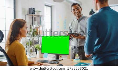 Three Start-up Entrepreneurs Work on Desktop Computer with Green Mock-up Screen, They Discuss Project Design, Use Touch Screen Digital Tablet. Modern Stylish Office with Diverse Team of Professionals.