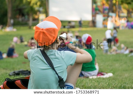 Open air cinema in the park. People sit in groups in isolation from each other. In the foreground, the boy sits turned to the screen.