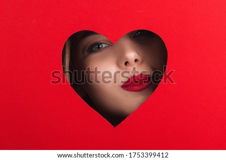 A beautiful girl with red lipstick, peeps out of a hole in shape of heart on red background. Commercial concept, postcard skit, store discount banner.