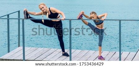 Sportive happy mother is doing workout with little daughter on the shore of the lake.