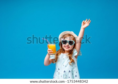 A cute little girl in a dress, hat and sunglasses poses on a pink background with a glass of cooling cocktail. child raises his hand up and rejoices at beginning summer holidays and goes on journey.