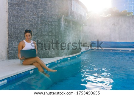 Stock photo of a caucasian woman sitting on the poolside with her feet in the water. She is using her laptop. She is wearing sunglasses.
