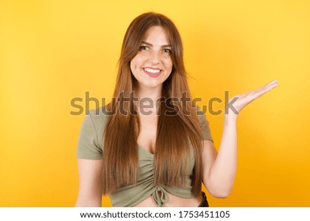Young caucasian woman with long hair wearing green tshirt standing over isolated yellow background smiling cheerful presenting and pointing with palm of hand looking at the camera.