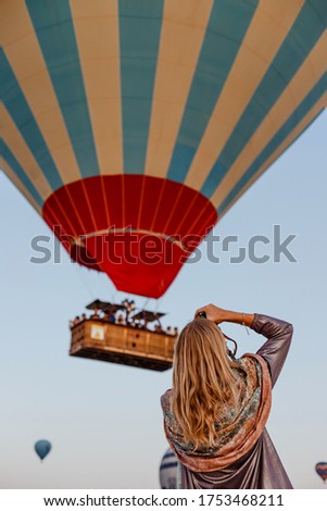 Beautiful blonde woman photographer takes pictures of flying hot air balloon at dawn in Cappadocia, Turkey. Travel concept.  Dressed in a scarf with a Turkish national pattern Ebru