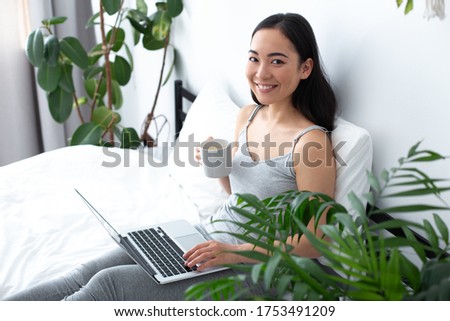 Happy pretty lady is sitting on bed with notebook on laps and enjoying hot drink in light room