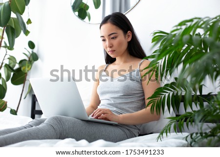 Young beautiful female is sitting on comfortable bed among green plants and typing on notebook