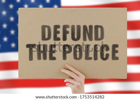 The phrase " Defund the police " on a banner in men's hand with blurred American flag on the background. Violence against black. Fight. Police reform. Occupation. Job. Protest. Brutality. Equality