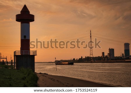 The picture shows a large river. A ship sails on the river which is illuminated by the sun. There is a lighthouse on the left.