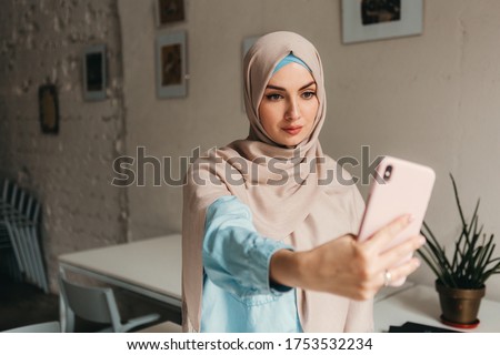 young pretty modern muslim woman in hijab working on laptop in office room, education online, remote work freelancer, taking selfie picture photo using phone camera