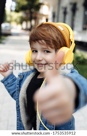 Photo of adorable little boy show thumb up listening music in headphones in street.