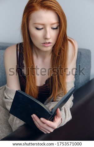 Red-haired girl sitting on the couch and looking at the black book in her hands.
