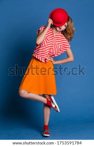 High fashion photo of a beautiful elegant young woman in a pretty  sleeveless red white shirt, orange skirt, socks, hat on blue background. Studio Shot.