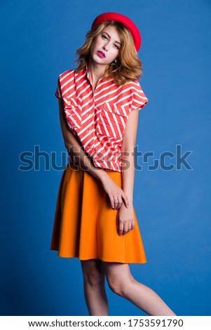 High fashion photo of a beautiful elegant young woman in a pretty  sleeveless red white shirt, orange skirt, socks, hat on blue background. Studio Shot.