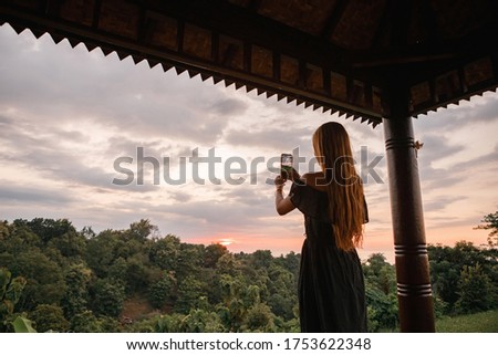 Back view young woman wearing dress stand on wooden terrace and taking picture on the phone, beatiful landscape wth greenery and sea view at sunset