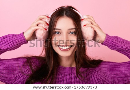 Cute Caucasian lady in a knitted sweater. She smiles broadly and holds fluffy pink winter headphones on her head. Photo on a pink background. Winter mood, winter clothes. New Year's bustle.