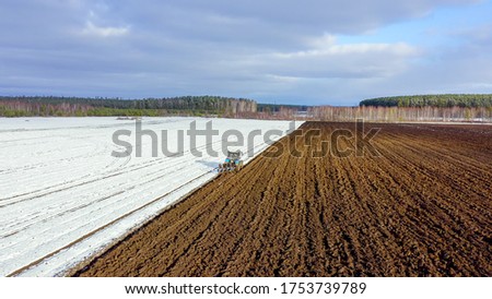 A blue tractor plows a field covered with snow. Behind the tractor is black earth. Russia, Ural, Aerial View  
