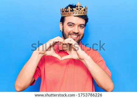 Young hispanic man wearing king crown smiling in love doing heart symbol shape with hands. romantic concept. 