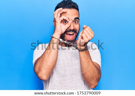 Young hispanic man wearing prisoner handcuffs smiling happy doing ok sign with hand on eye looking through fingers 