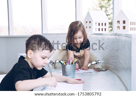 Little girl in a full skirt draws with crayons. In the foreground her brother is in defocus.