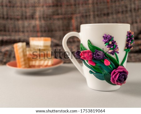 The original mug with decorative figurines made with their own hands. Figures on the cup are fashioned hand made from colored clay. Plate of sweets on a background. High quality photo