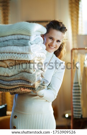 smiling elegant woman in white sweater and skirt in the modern house in sunny winter day looking out from huge pile of sweaters.