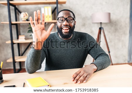 Excited African-American young guy in glasses looks into camera and greeting , waving hello. Video screen, video chat, online call, webcam shot