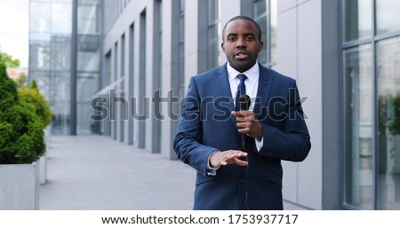 Portrait shot of young African American handsome male journalist talking with microphone for news episode outdoor. Pandemic concept. Man correspondent in suit and tie and with mic. Royalty-Free Stock Photo #1753937717