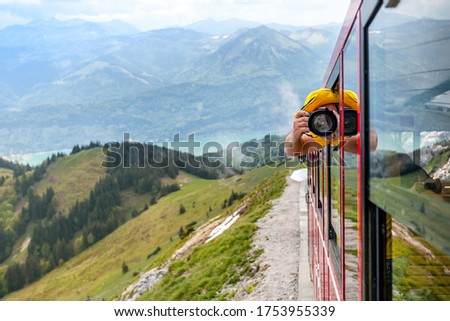 A man in yellow clothes photographs leaning out of a train window on Schafberg mountain, Austria.
