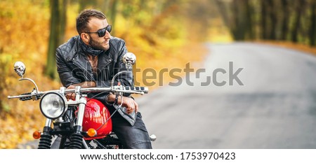 Bearded brutal man in sunglasses and leather jacket sitting on a motorcycle on the road in the forest with blured colorful background.