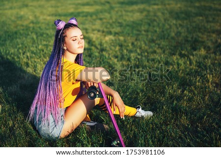 Young beautiful girl sitting on a lawn holding a skateboard in her hand in a yellow T-shirt and yellow socks in the open.