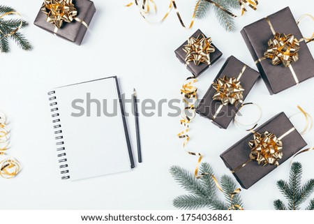 Festive Christmas flat lay blank notebook next to fir branches and black gift boxes with golden bows on white background.