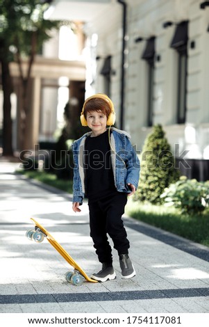 Photo of joyful little boy posing with headphones and skateboard in street.