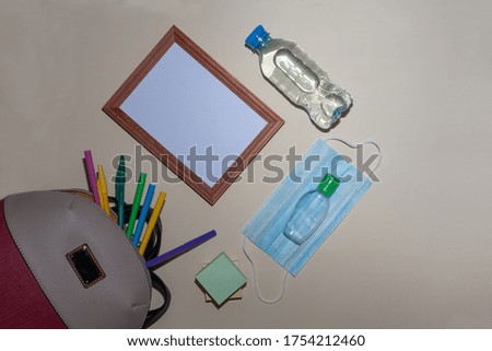There is a sanitizer on the anti-virus mask. Photo frame and a bottle of water. Colored pens poke out of the rucksack and there is a block for writing on them.