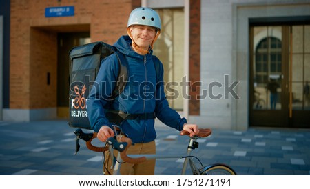 Handsome Happy Food Delivery Man Wearing Thermal Backpack and Safety Helmet Stands Beside his Bike in the Stylish Modern City District. Portrait of the Smiling Courier Delivering Restaurant Order