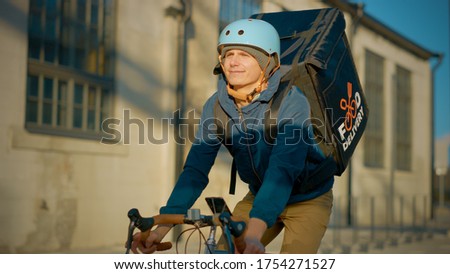 Food Delivery Courier Wearing Thermal Backpack Rides a Bike on the Road To Deliver Orders for Clients and Customers. Sunny Day in Modern City with Stylish Buildings.