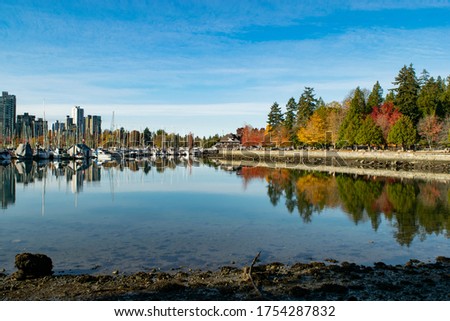Skyline with city view and port to the left and park trees in different colors to the right. Sunny day with blue sky and the reflection of the subjects is clear to see on the calm water of the river.