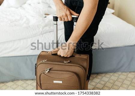 Young man raises the handle of a suitcase on wheels in a hotel. Departing for a trip with brown carry-on baggage. Guy takes brown suitcase on wheels. blurred baggage in motion