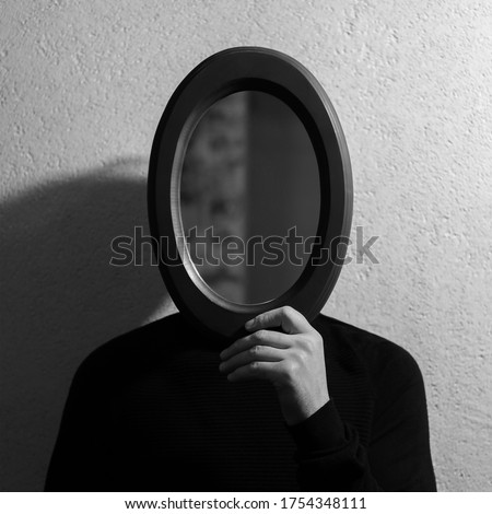 Black and white studio portrait of young man holding oval mirror on face. Background of textured wall.