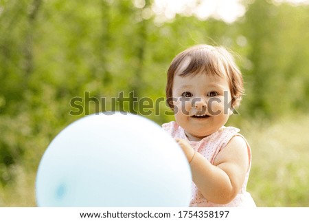 Cute, smiling child holds a bright balloon. Outdoors. Green trees and grass on the background. Summer. Caucasian. Positive emotions, happy baby. Copy space. Lifestyle. Eye contact. Soft focus. 