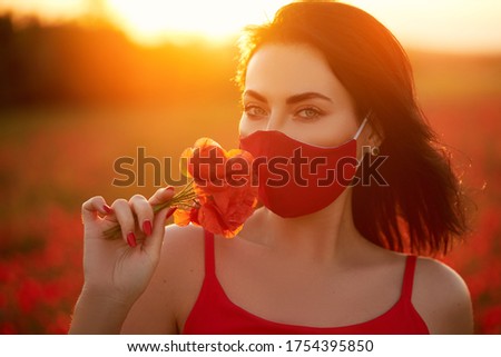 Fashion quarantine look. Young woman model in red antiviral handmade mask sniffing flowers on the background of a sunset in a field of red poppies. Summer fashion