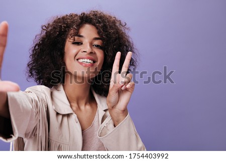 Brown-eyed woman in beige outfit shows peace sign and makes selfie. Charming girl in jacket widely smiling on purple backdrop