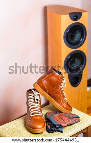 Closeup of Protective Motorcyclist Leather Tan Sneakers Placed Indoors Along With Leather Gloves Against Floorstanding Loundspeaker. Vertical Image composition