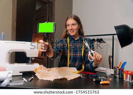 A girl seamstress keeps a blog about sewing in a workshop at home. Craftsman holds smartphone with green screen.